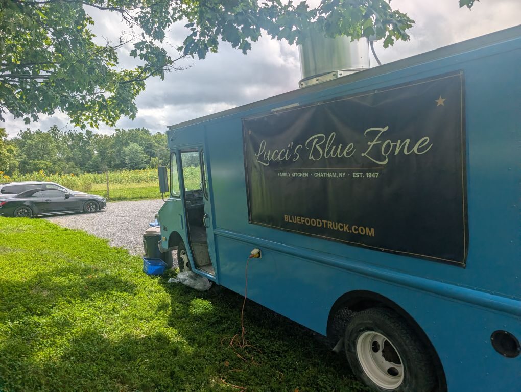 Lucci's Blue Zone truck parked under the trees at Chatham Berry Farm with wildflower field behind